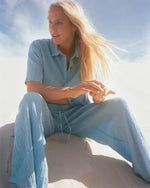 Woman wearing a light blue short-sleeve button-up shirt with matching textured pants, seated outdoors.