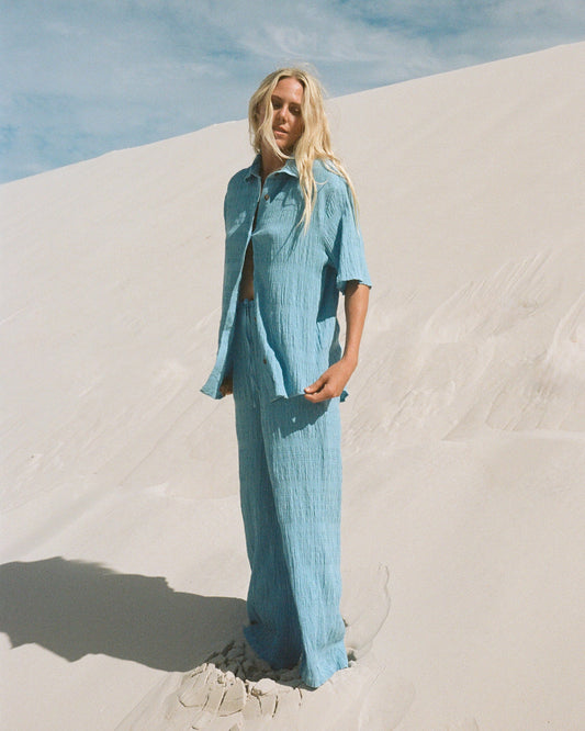 Woman wearing blue elastic waist pants with matching button-up shirt in textured fabric, styled on white sand.