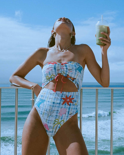 White bandeau one-piece swimsuit with blue gingham pattern and multicolored beach-themed graphics including palm trees and st
