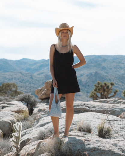 Black sleeveless slip dress with thin straps worn with a straw cowboy hat and blue shoulder bag.