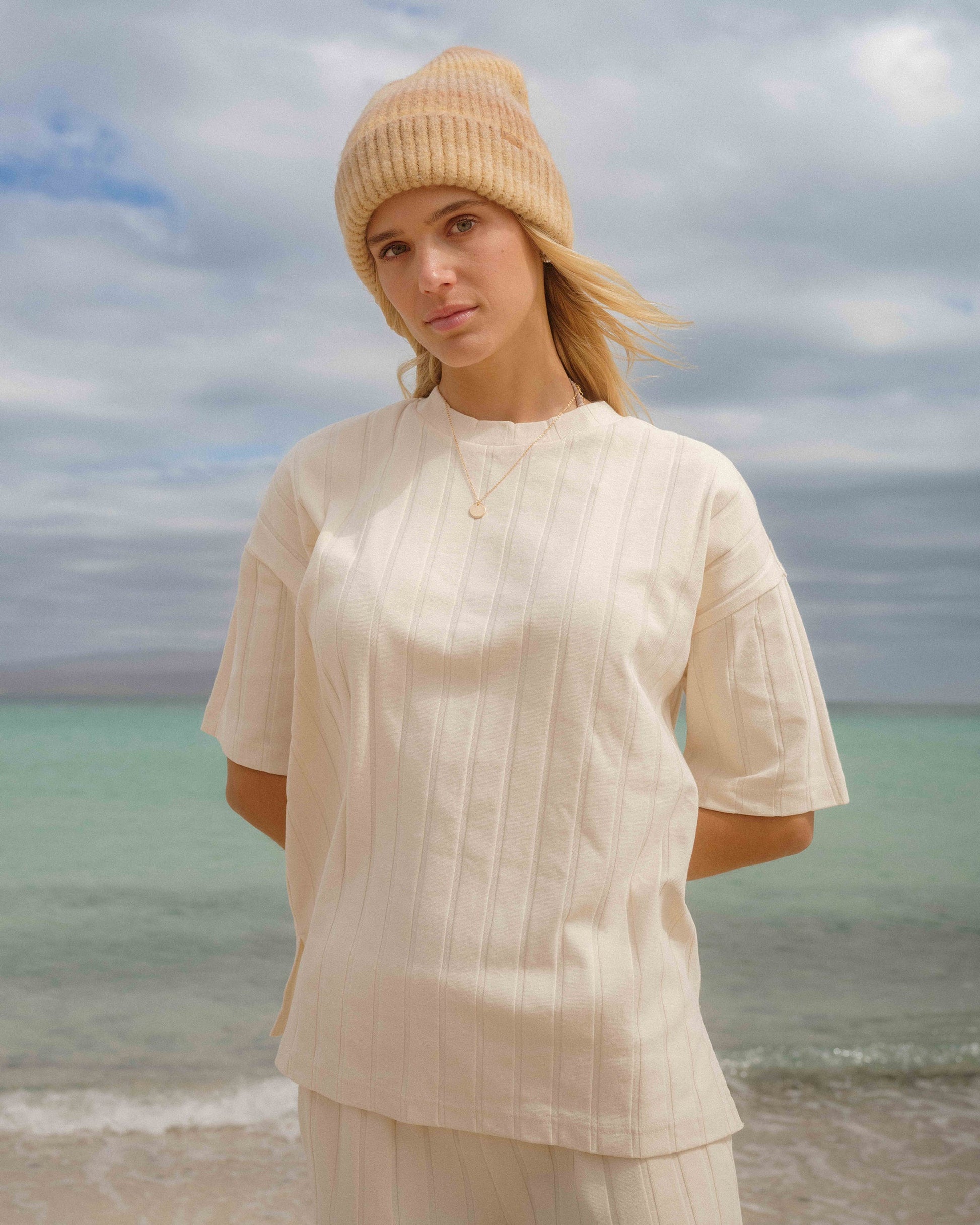 White ribbed short-sleeve top with vertical texture detail worn with a cream beanie by the ocean.