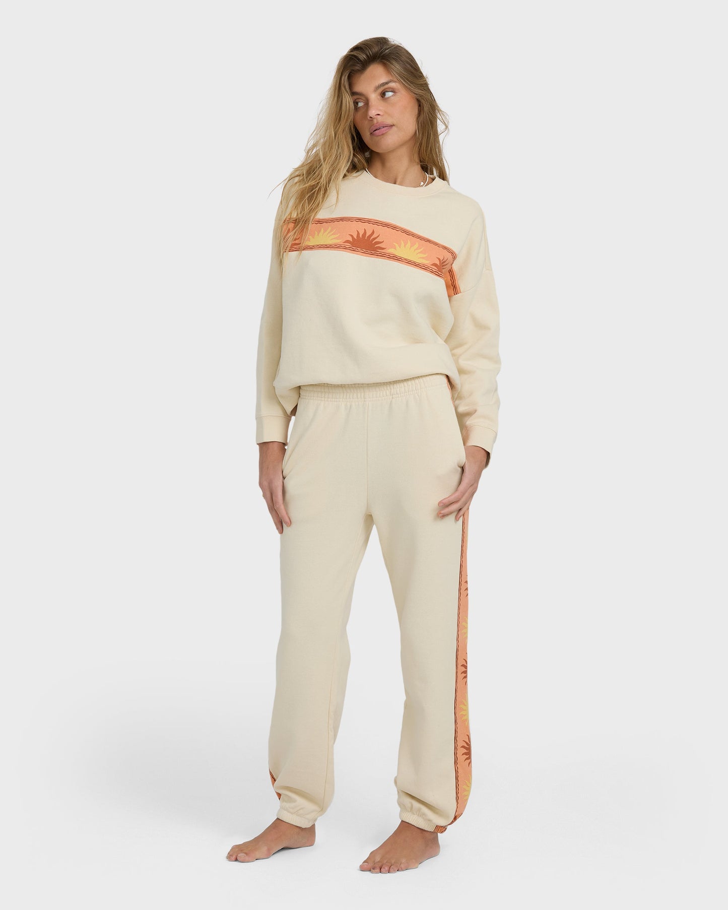 Woman wearing cream Echo Jogger set with orange sun-print stripe detail on crewneck sweatshirt and jogger pants.
