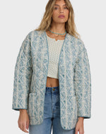 Mineral blue quilted jacket with geometric pattern, worn open over a patterned top and jeans.