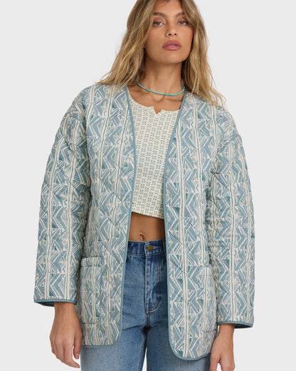 Mineral blue quilted jacket with geometric pattern, worn open over a patterned top and jeans.