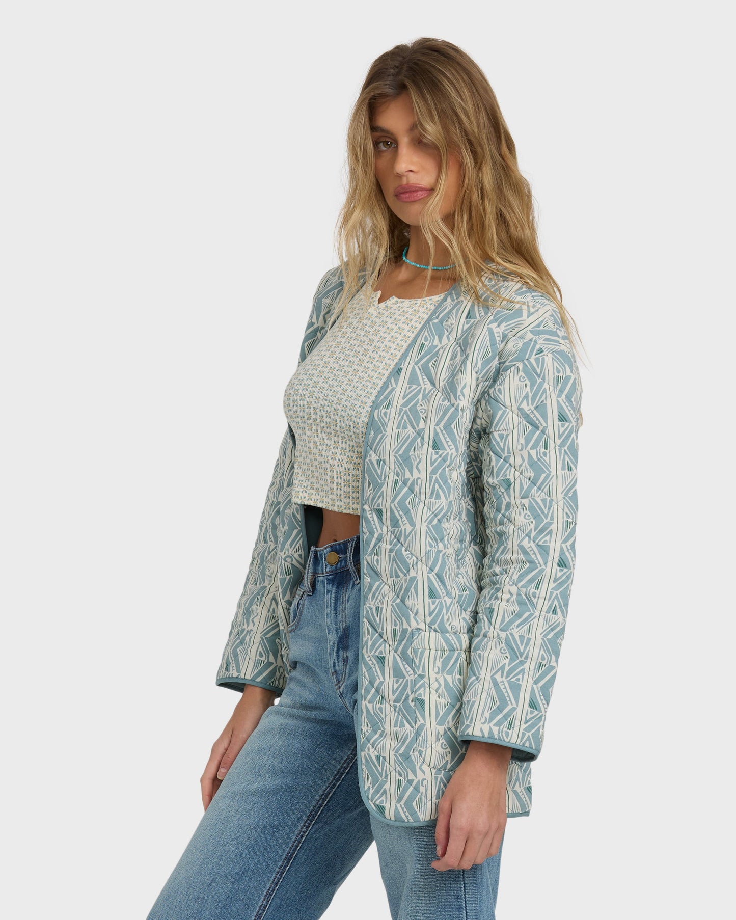 Mineral blue quilted jacket with geometric pattern worn over a cream textured top and blue jeans.