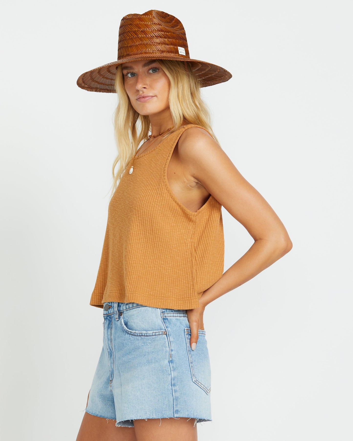 Woman wearing light blue denim shorts with raw hem, tan ribbed sleeveless tank top, and brown woven straw hat.