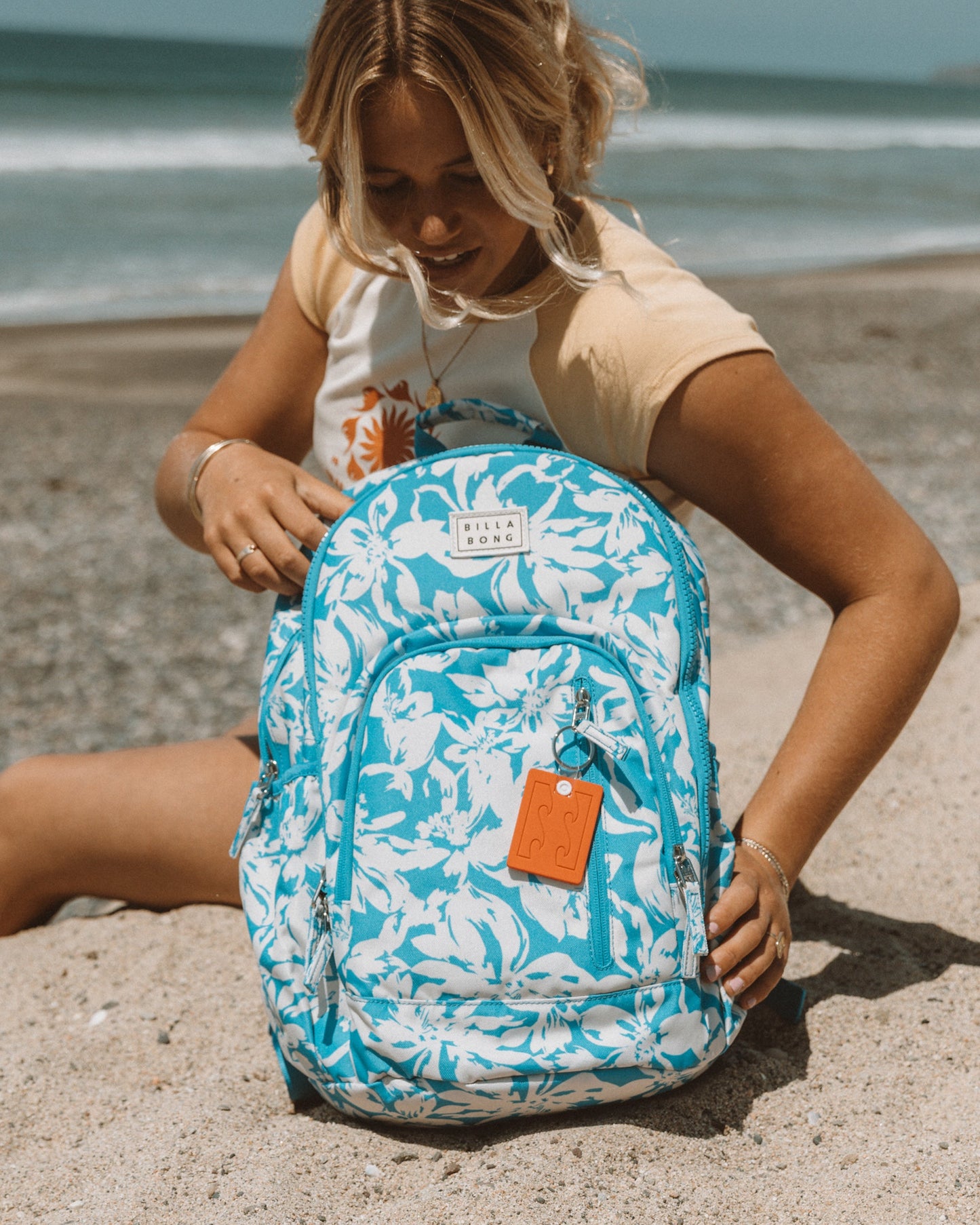 Blue and white floral backpack with multiple compartments and orange keychain attachment, displayed on beach sand.