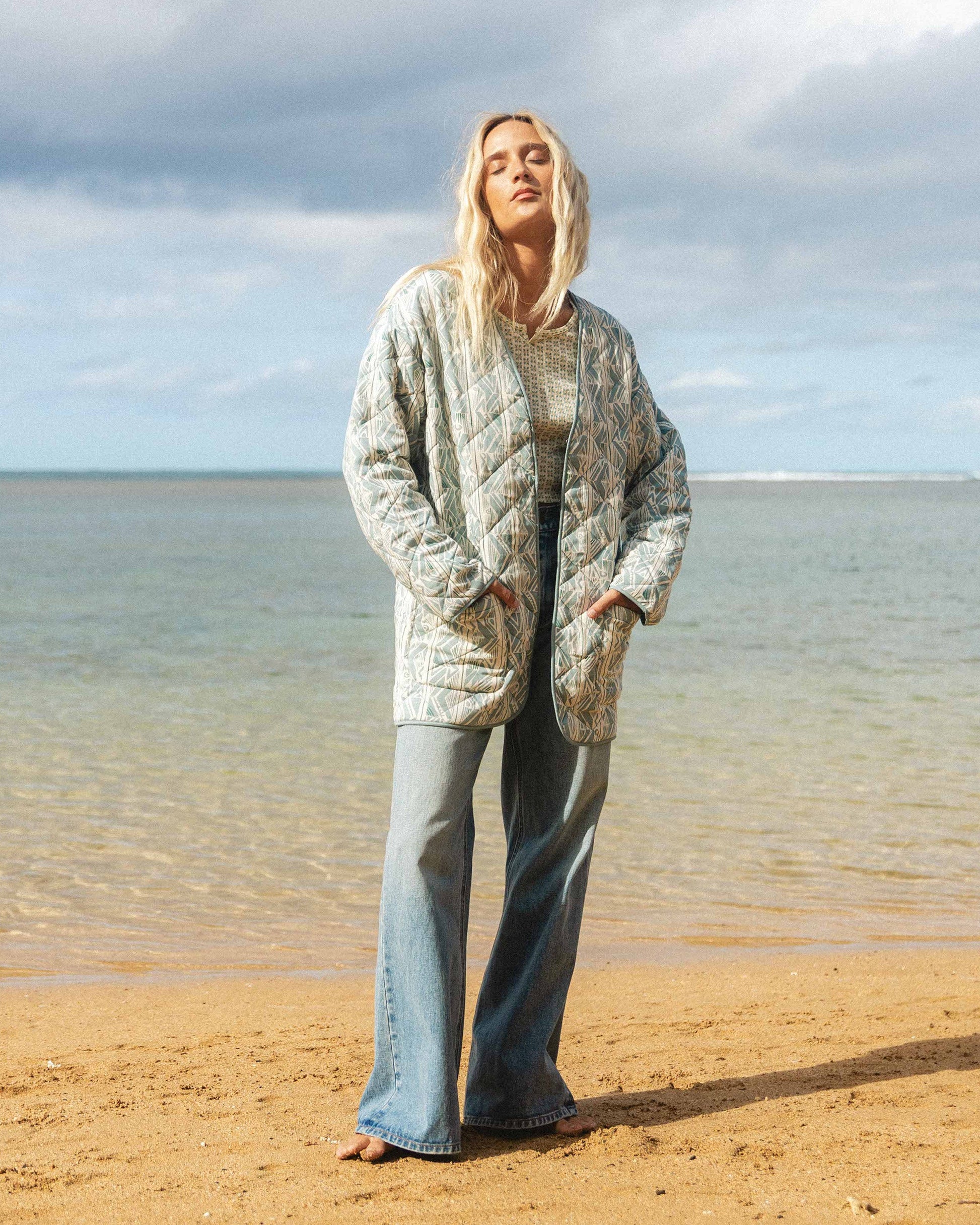 Woman wearing a mineral blue quilted jacket with geometric pattern over a knit top and jeans at the beach.