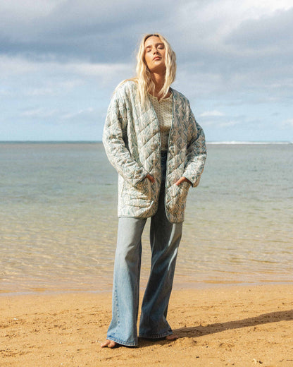 Woman wearing a mineral blue quilted jacket with geometric pattern over a knit top and jeans at the beach.