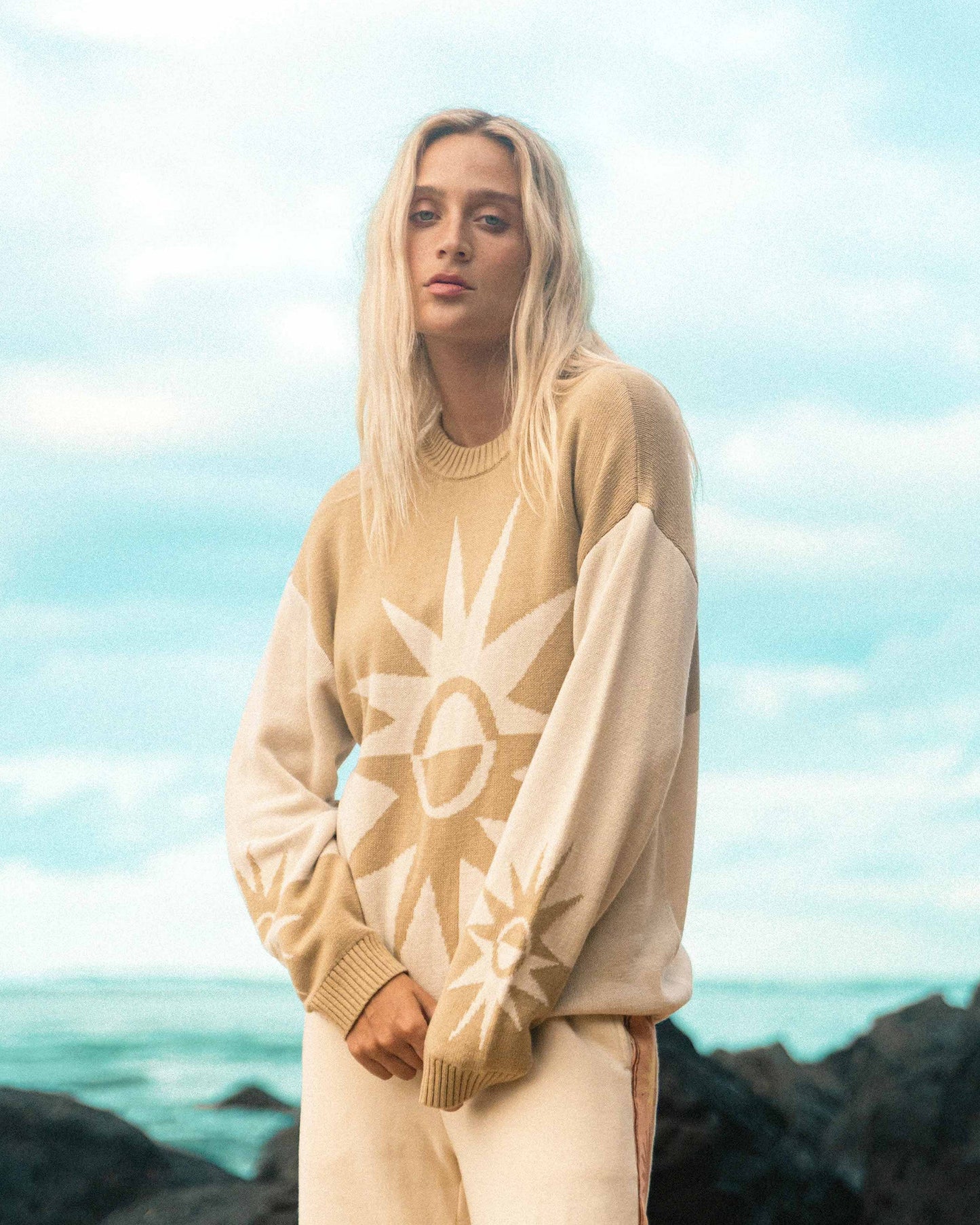 Cream-colored sweater with sun graphic design and ribbed collar and cuffs worn by person against ocean background.