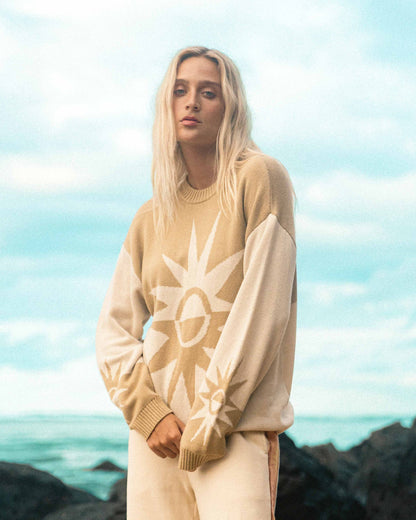 Cream-colored sweater with sun graphic design and ribbed collar and cuffs worn by person against ocean background.