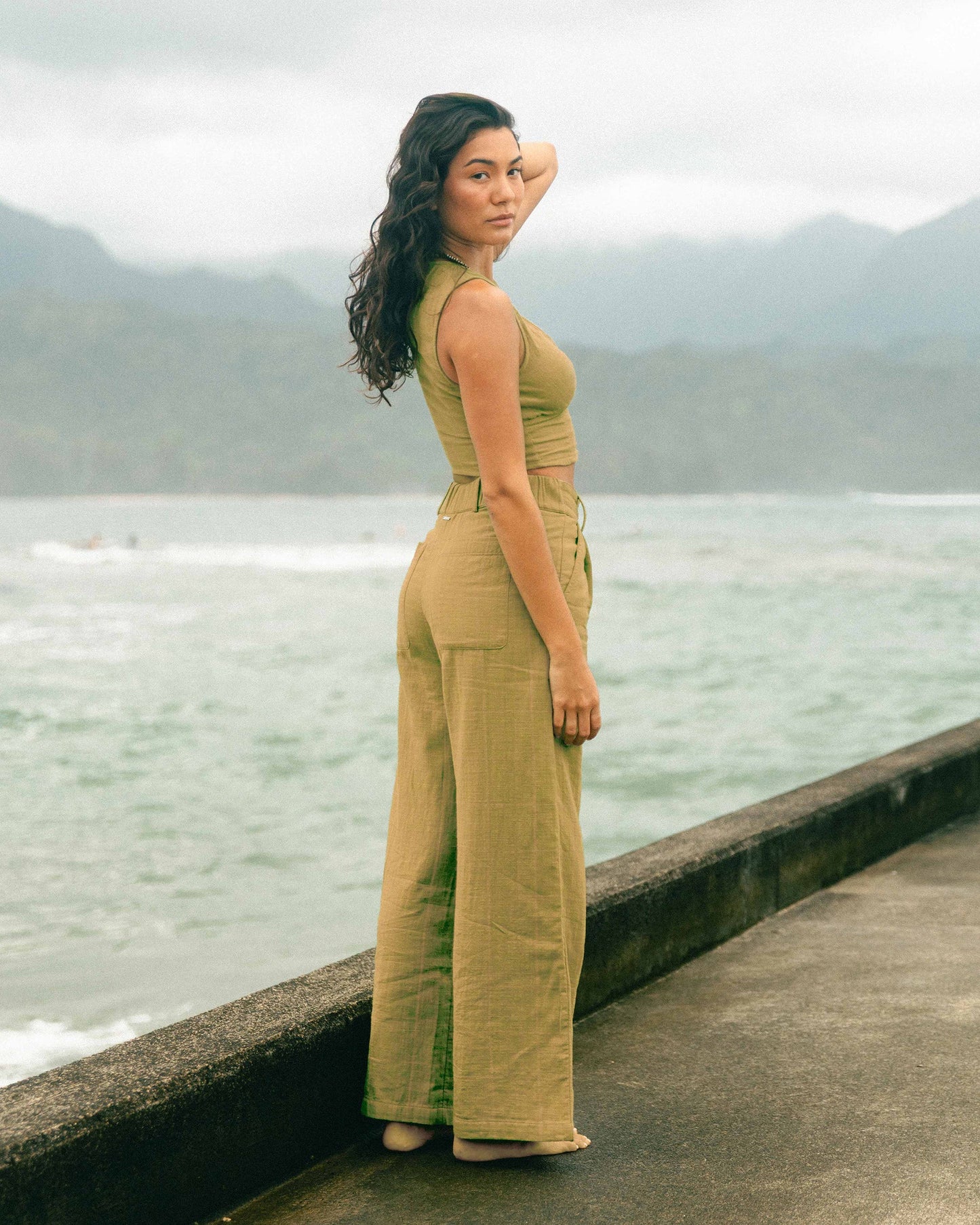 Woman wearing mustard-yellow high-waisted wide-leg pants with matching sleeveless top, standing by water with mountains in ba