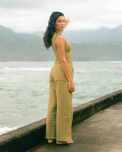 Woman wearing mustard-yellow high-waisted wide-leg pants with matching sleeveless top, standing by water with mountains in ba