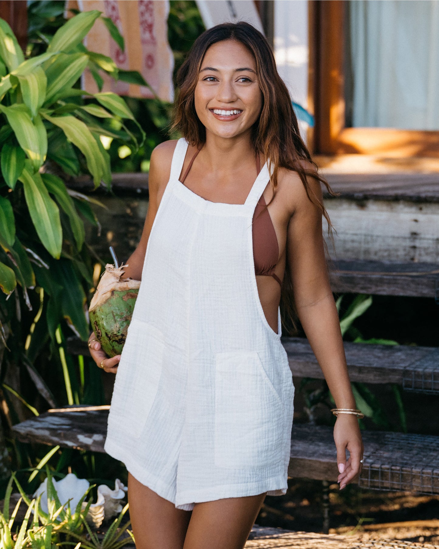 Woman wearing white sleeveless romper with square neckline, standing outdoors among tropical plants.