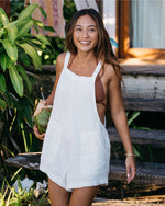 Woman wearing white sleeveless romper with square neckline, standing outdoors among tropical plants.