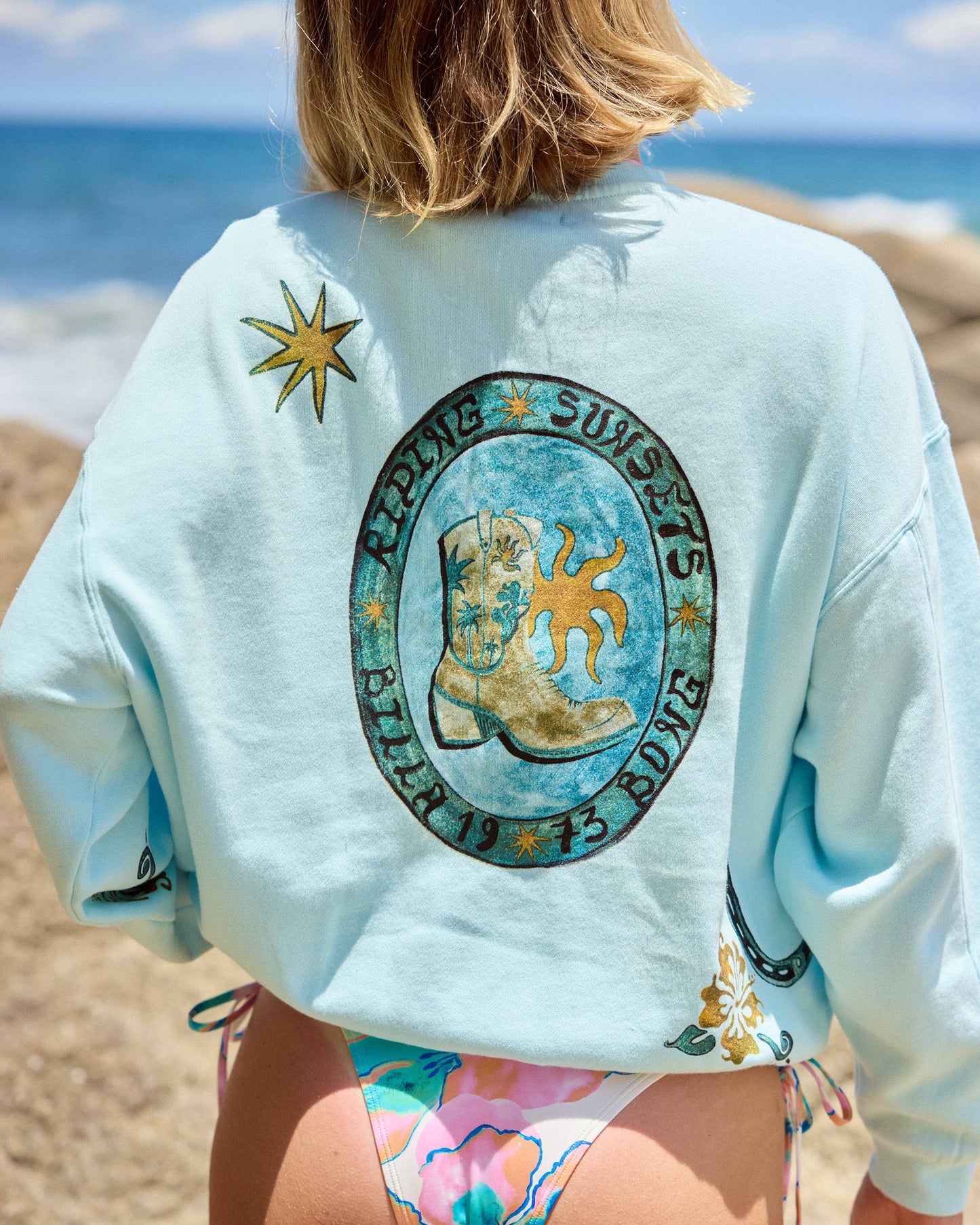 Clearwater crewneck sweatshirt with sun and surfboard graphic on back, worn at beach.