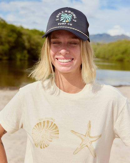 White short sleeve t-shirt with gold shell and starfish graphics, styled with a navy baseball cap.
