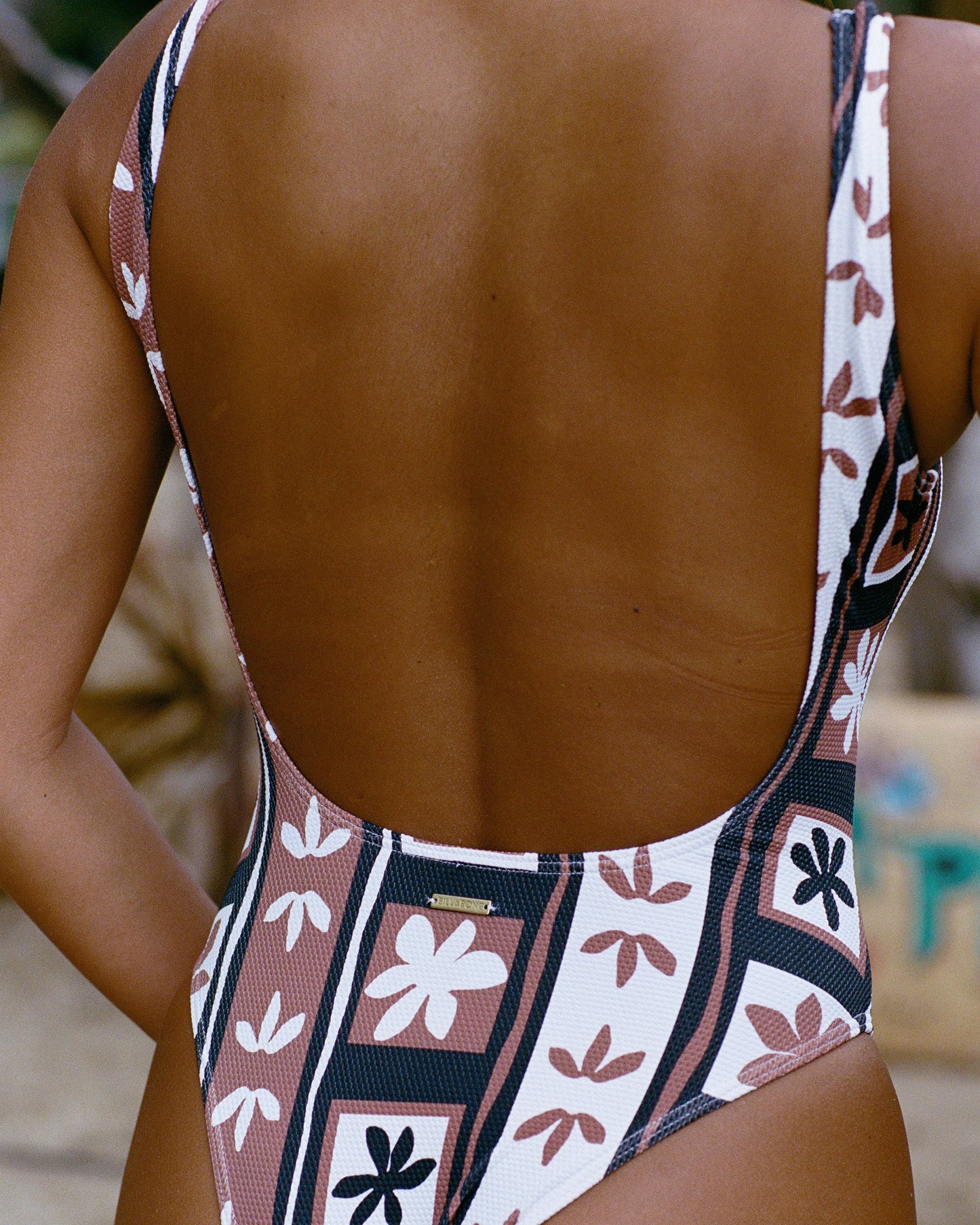 Brown stone one-piece swimsuit with open back and floral patterned straps in cream, brown, and navy colors.