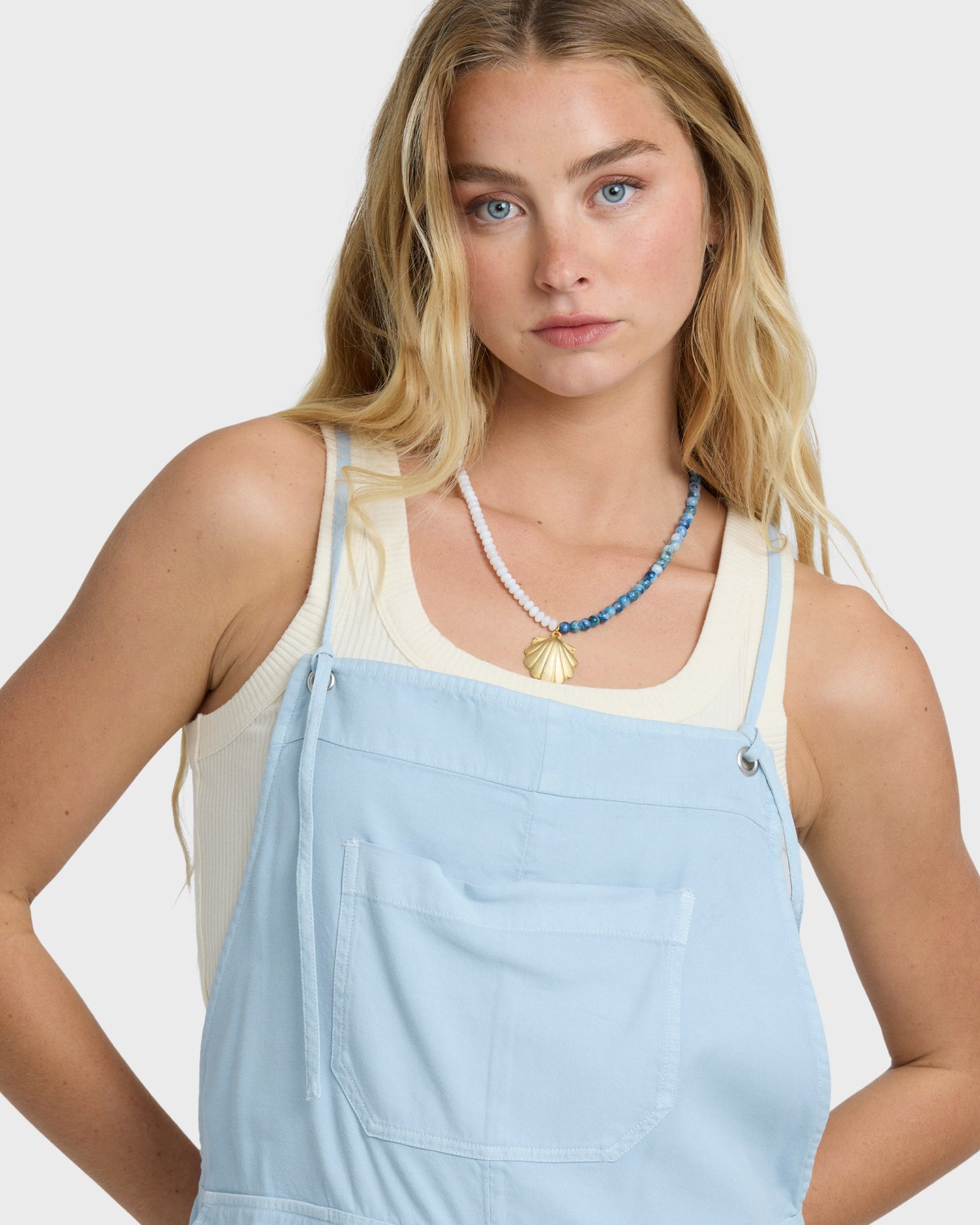 Woman wearing light blue chambray overalls with a cream tank top and beaded necklace.