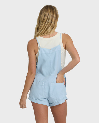 Woman wearing light blue chambray overalls with rolled shorts hem and a white ribbed tank top, shown from back.