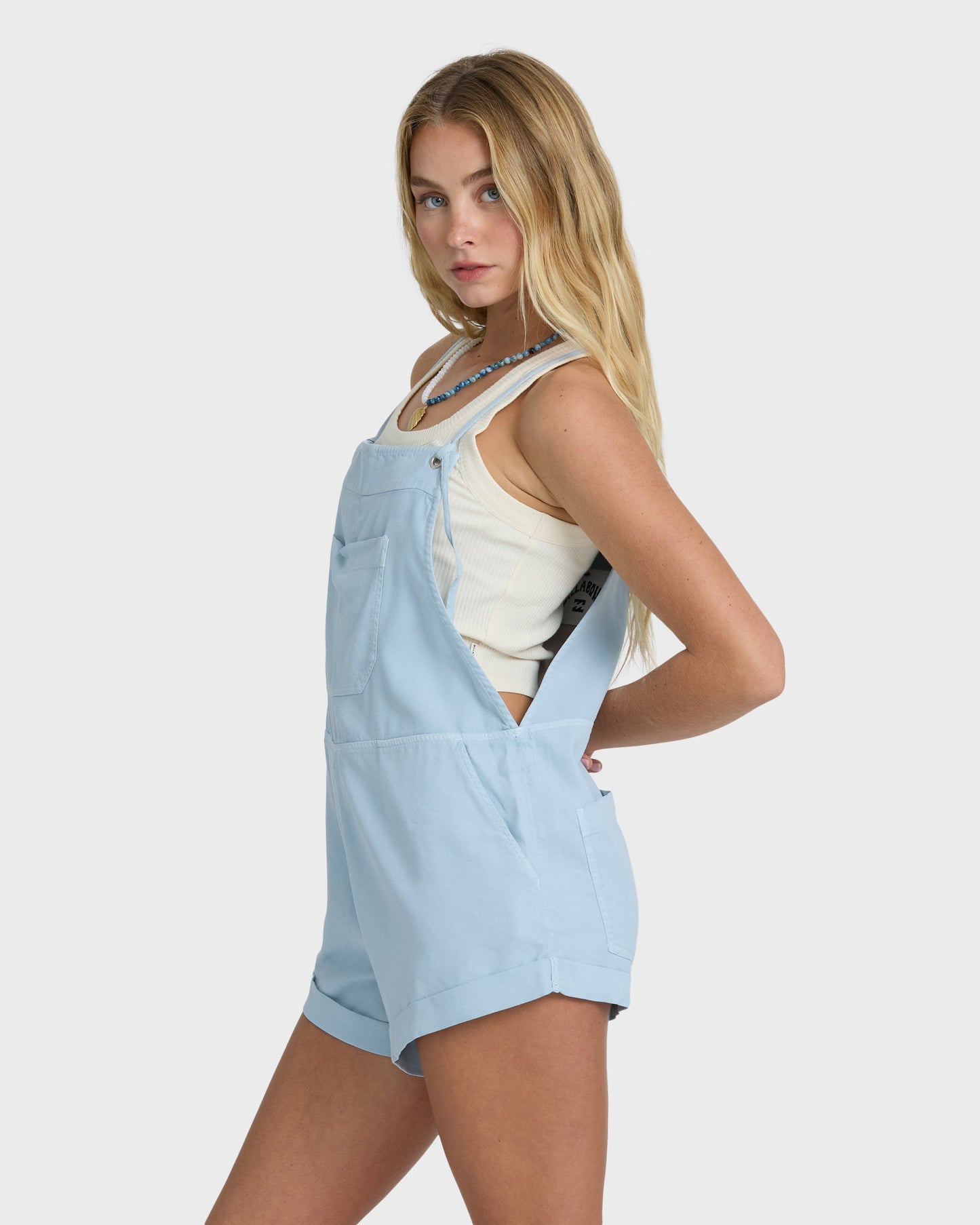 Light blue washed chambray overalls with front pockets and adjustable straps, worn over a white tank top.