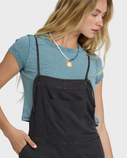 Woman wearing off-black yarn-dyed overalls with adjustable straps over a blue t-shirt.