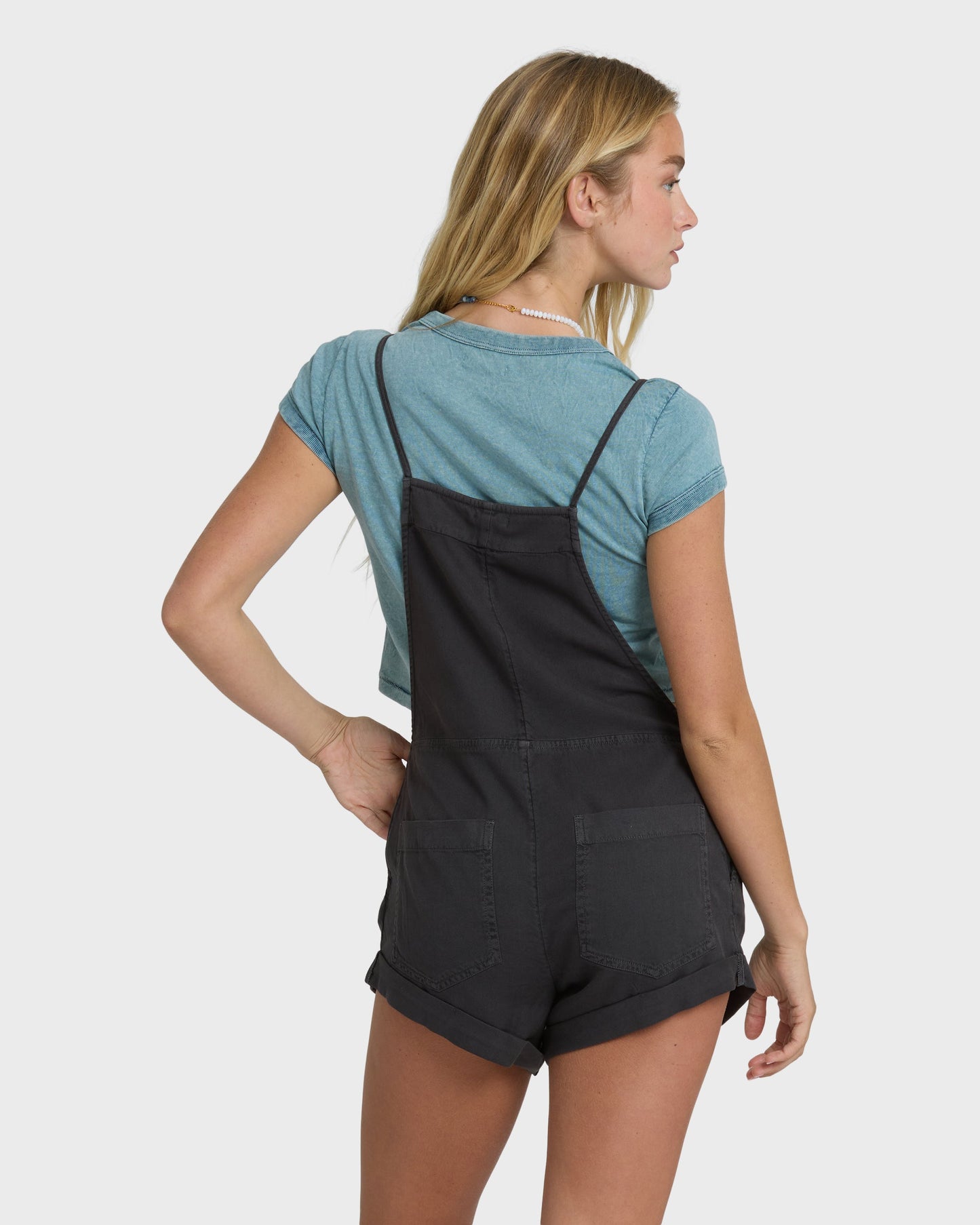 Woman wearing off-black denim overalls with adjustable straps over a blue t-shirt, shown from the back.