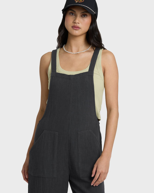 Black sleeveless jumpsuit with square neckline and front pockets, styled with cream ribbed tank underneath.