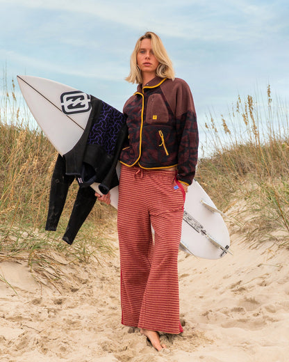Woman wearing rose brown horizontal striped terry pants with yellow trim at beach, holding surfboards.